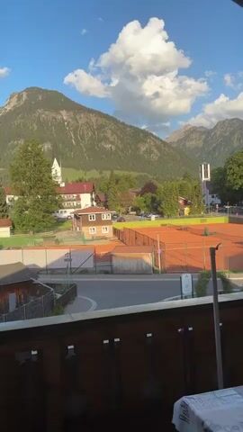 The video shows a view from a balcony overlooking a scenic town nestled in front of mountains. The foreground features a tennis court with red clay, separated from the viewer by a road and a low fence. Beyond the tennis court, there are various buildings including a church with a tall steeple, residential houses with red roofs, and several trees. The background is dominated by a large, forested mountain with a few scattered clouds in the blue sky. The view is framed by the balcony's wooden railing.