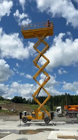A person wearing a hard hat and a safety vest stands on a raised platform of a yellow scissor lift. The scissor lift is fully extended, reaching a significant height against a blue sky with white clouds. The lift is situated on a paved surface, possibly a construction site, with unfinished concrete structures visible in the foreground. In the background, a truck with an orange trailer is parked, and there are trees and a hillside. The video captures the stationary scissor lift with the person on top.