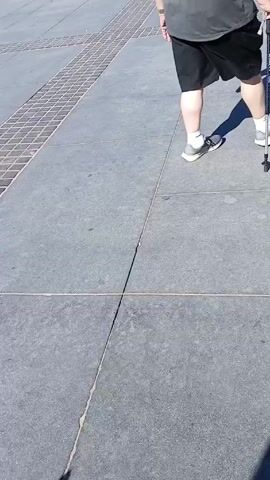 The video shows a sunny day at a bustling outdoor pedestrian area. A person with a walking stick is walking through the crowd, heading towards a street lined with palm trees and buildings. There are many people walking around, and outdoor seating areas for restaurants can be seen. The ground is paved with grey stone, with a grid pattern of darker stones. The sky is clear and blue.