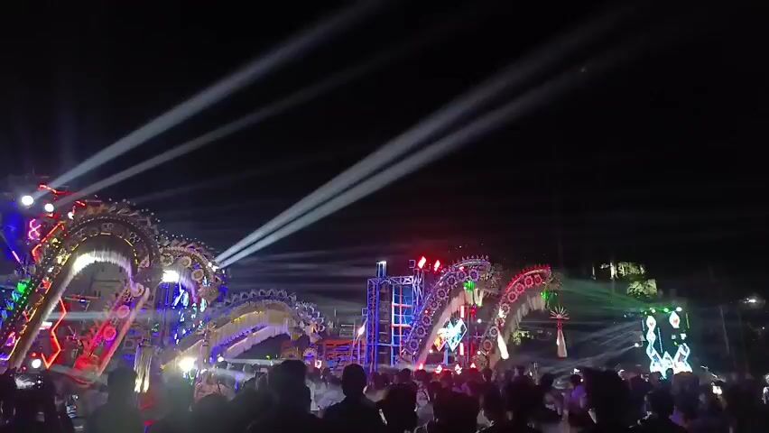 The video shows a nighttime outdoor concert or festival with a large crowd in the foreground, silhouetted against a brightly lit stage. The stage features elaborate, dragon-like structures adorned with colorful lights, including red, green, blue, and gold. Powerful spotlights are cutting through the dark sky from behind the stage. The crowd appears to be actively engaged, with many people facing the stage and some raising their hands. The atmosphere is energetic and festive.