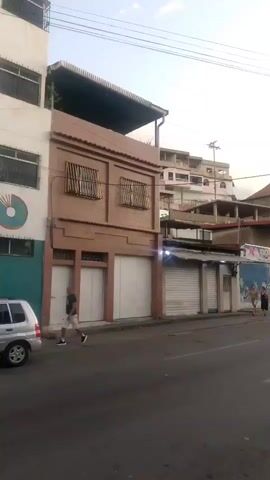 The video pans across a street scene. On the left, there is a two-story brick building with closed storefronts on the ground level. Above the storefronts, there are windows with metal bars. A man in a dark shirt and shorts walks on the sidewalk in front of this building. Further down the street, there are more buildings with various architectural styles, some appearing to be multi-story residential buildings on a hillside. Several storefronts with rolled-down metal shutters are visible. Two women are standing on the sidewalk in front of one of these shops. A silver car is driving on the street. In the distance, there is a tree and more buildings, and the sky is overcast. The overall impression is a street in a residential or mixed-use urban area, possibly in a developing country, during daylight hours.