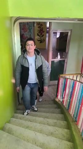 The video shows a group of young people walking down a set of stairs inside a building. The walls are painted a bright green. The stairway has a colorful railing with vertical bars of different colors. Several individuals are walking down the stairs, and they appear to be in casual winter clothing. The environment looks like it could be inside a school or community center. The light is bright and seems to be coming from natural sunlight.