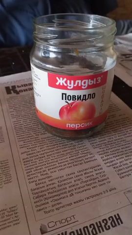 A glass jar of peach jam sits on a newspaper on a table. The jar is about three-quarters full. The label on the jar is red and white with an image of two peaches. The text on the label is in Cyrillic. The newspaper is open to an article with a headline in large, bold print. The background is blurred but appears to be a room with furniture. The lighting is natural, coming from a window or light source to the left of the frame. There is no visible person in the video. The scene is static.