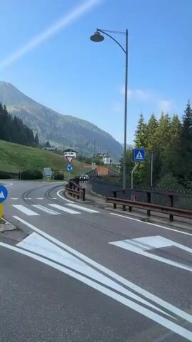 The video shows a view from a moving vehicle on a paved road. The road has white lane markings. On the right side of the road is a metal guardrail with a wooden top rail. Beyond the guardrail, there is a grassy area with a small body of water and some trees. In the background, there are mountains covered in trees and some houses. The sky is blue with some wispy clouds. There is a street light and road signs visible on the left side of the road at the beginning of the video. The video appears to be recorded during the daytime, likely in the morning or afternoon.