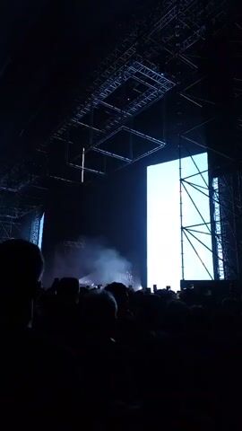 A crowd of people is at a concert. The stage is lit with blue spotlights and has large screens showing images of the band members. The stage has metal trussing around the screens. There is a drum set visible on the stage. The audience is holding up their phones to record the performance. There is smoke and light on the stage.