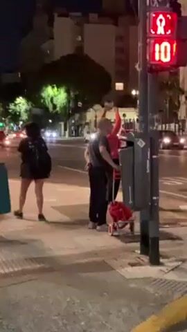 The video shows a nighttime street scene with several people gathered on a sidewalk in an urban environment. A traffic light is visible in the foreground with a countdown timer displaying the number "10". People are standing and walking around, some with bags or small wheeled carts. There is a stack of wooden planks or boards near the sidewalk, with several people standing or sitting on top of them. The people are diverse in age and clothing. The scene appears to be at a street corner, with buildings and streetlights visible in the background. Cars pass on the street. The scene appears to be a gathering or event of some kind.