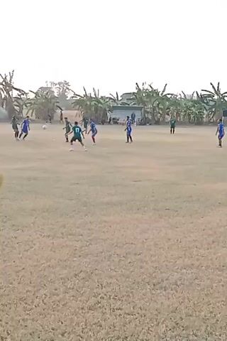 A soccer game is underway on a dry, grassy field. The sky is bright white. Several players in blue and green uniforms are scattered across the field, some with their backs to the camera. One player in a blue uniform kicks a white soccer ball. The players appear to be young men. In the background, there are trees, likely banana trees, and a few buildings. The scene is static with minimal movement.