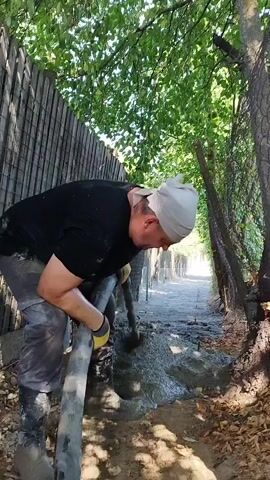A man wearing a black t-shirt, gray work pants, and black boots, with a white cloth wrapped around his head, is holding a concrete hose with yellow and black gloves on his hands. He is directing the hose towards the ground, where wet concrete is being poured. The man is positioned on the left side of the frame, leaning forward and concentrating on his task. The environment appears to be outdoors, with a dirt path lined by a fence on the left and trees on the right. The background shows a continuation of the path, also lined with vegetation. The lighting suggests it is daytime.