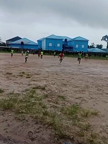 A group of young people in athletic attire are playing soccer on a muddy, uneven outdoor field. Several individuals are visible in motion, kicking and running with a soccer ball that is not clearly discernible. The environment is overcast, suggesting cloudy or rainy weather, with low-lying buildings in the background. The field is mostly bare earth with patches of sparse grass and some puddles of water. The scene appears to be a casual game or practice session.