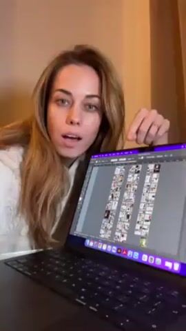 A woman with long brown hair and blue eyes is holding a laptop computer. The laptop screen displays a grid of thumbnail images, arranged in rows and columns, suggesting the layout of a multi-page document, possibly a magazine. The woman is looking directly at the camera, pointing at the screen, and speaking. She says she created an 88-page magazine. She is wearing a light-colored hooded sweatshirt. The background is a light beige wall with a soft glow.