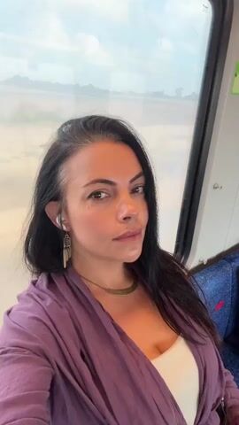 A woman with dark hair and fair skin is sitting on a train, looking directly at the camera. She is wearing a purple shawl over a white top and a gold necklace. Her hair is long and dark, pulled back slightly from her face, revealing earrings with a feather-like design. A faint smudge of makeup is visible on her cheek, possibly from the reflection of her hand. The train window behind her shows a blurred landscape of fields and trees under a cloudy sky. The interior of the train is visible, with blue seats and a portion of a metal pole. The lighting is natural, coming from the window. The video is short and seems to be a selfie taken while traveling.