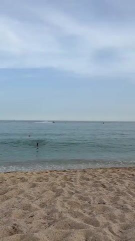 The video shows a beach with sand in the foreground and the ocean in the background. The sky is cloudy. A bird is flying in the sky. There are a few people in the water and a jet ski in the distance.
