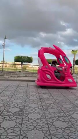 A small pink electric toy car slowly turns in a paved park area. The car has an open cockpit and the suggestion of aerodynamic styling. A child appears to be driving. The park has trimmed hedges and palm trees. In the background, several apartment buildings are visible under a partly cloudy sky. A streetlight stands nearby. A car moves on a road behind a low fence.
