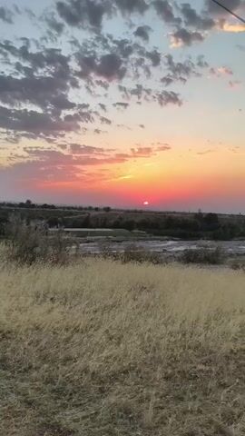 The video captures a sunset over a dry, grassy plain with a river in the middle ground. The sky is filled with dramatic clouds illuminated by the setting sun, casting hues of orange, pink, and yellow. The foreground is dominated by tall, dry grass, and the background shows a treeline and a hint of distant landscape. The scene is peaceful and natural, with no people or man-made objects clearly visible.