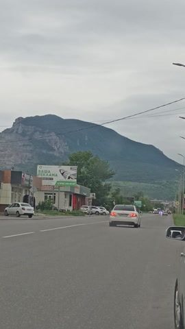 The video shows a road passing through a town with mountains in the background. Cars are driving on the road, and there are buildings and billboards on the side. The sky is cloudy.