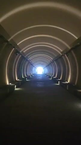 This is a first-person perspective video of a person walking through a dimly lit, circular tunnel. The tunnel is illuminated by lights positioned along the walls and at the end of the tunnel where there is a bright light source. The walls of the tunnel have a reflective quality that bounces the light, creating concentric circles of light on the ceiling. The person is walking on a paved surface, and there are railings on either side of the path. The person's feet are visible at the bottom of the frame. The video captures the sound of footsteps and what appears to be faint vocalizations or exclamations.