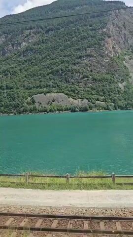 The video shows a moving train track in the foreground with gravel and grass. Beyond the tracks, there is a turquoise lake with a forested mountain rising steeply behind it. The sky above is cloudy. The video is shot from the perspective of someone on a moving train.