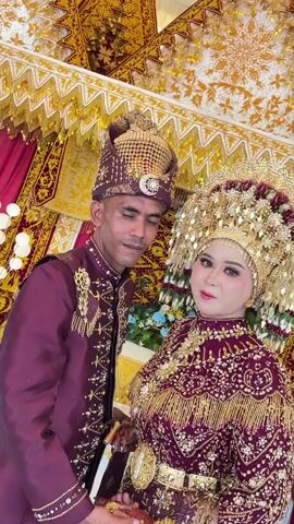 The video shows a wedding ceremony. A man and a woman, dressed in traditional wedding attire, pose for a photo. The man is wearing a maroon suit with gold embroidery and a matching hat. The woman is wearing a maroon dress with gold embellishments and a large, ornate gold headdress. They are standing in front of a backdrop decorated with gold and maroon fabric. The man holds a bottle in his left hand and waves with his right hand. The woman also waves with both hands. The couple smiles at the camera.