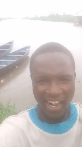 This is a selfie video taken from the chest up of a young Black male. He is smiling and facing the camera. He is wearing a white crewneck sweatshirt with a light blue collar. Behind him, there is a body of water with two canoes floating on the left side of the frame. In the distance, on the right side of the frame, there is a structure that appears to be a house or building on stilts over the water. The sky is overcast and hazy, and the overall lighting is diffused. The environment appears to be a waterfront or riverside setting.