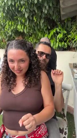 A medium shot shows a woman in a brown tank top dancing with her arms and fists clenched. A man in a black polo shirt and sunglasses is standing behind her with his arms around her. They are outside, with a hedge behind them and other people in the background. The woman has curly hair, a red bandana around her waist, and a wedding ring. The man has a beard. The woman is looking at the camera and smiling. The man is looking around and occasionally looking at the camera, making faces. There is music playing in the background.