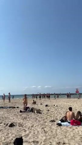 The video shows a crowded beach on a sunny day. A large group of people stands in the ocean, tossing a ball. In the foreground, several individuals are lying on the beach, relaxing. The sand is light-colored. The sky is clear and blue. There is a structure, possibly a pier or breakwater, visible in the distance. A red sign is mounted on it. Beachgoers have personal belongings scattered around them.