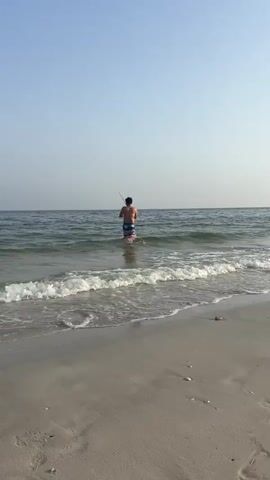 A man in shorts stands in the ocean with a fishing pole. A fishing rod holder is placed in the sand at the edge of the water. The waves are gently rolling onto the sandy beach. The sky is clear and blue.