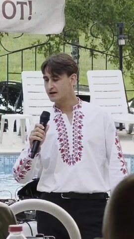 A young man stands in front of a body of water, holding a microphone and singing. He is wearing a white shirt with red embroidery on the collar and sleeves, and black pants. In the background, there are white lounge chairs and trees. A banner with some text on it is visible at the top of the frame. There are also some people in the foreground, out of focus. The overall scene appears to be an outdoor event.