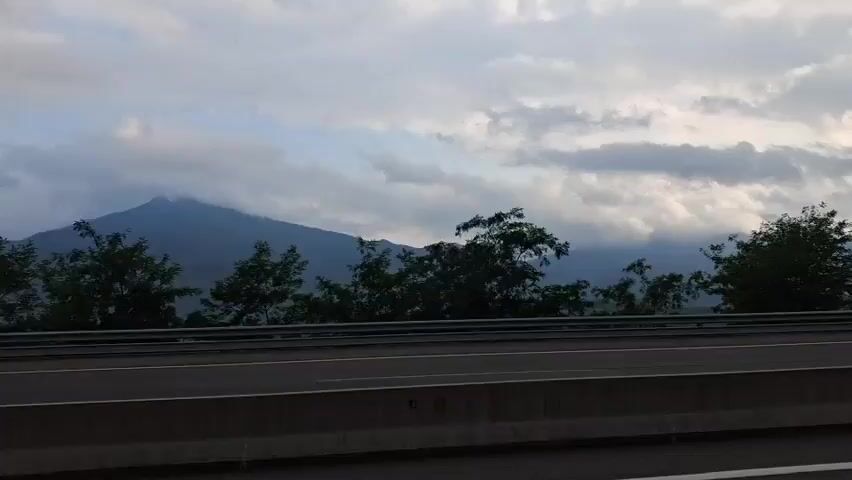 The video captures a scenic view from a vehicle traveling on a highway. The primary subject is the expansive landscape of mountains and trees, with a cloudy sky overhead. The highway itself, with its concrete barriers and lane markings, occupies the foreground. In the right side of the frame, a side-view mirror reflects the road ahead, indicating the direction of travel.

The scene appears to be set in a mountainous region, suggested by the rolling hills and peaks in the background. The presence of trees, some of which are quite dense, further emphasizes the natural setting. The weather seems overcast, with significant cloud cover obscuring parts of the mountains, lending a somewhat dramatic or moody atmosphere to the scene. The time of day is difficult to pinpoint precisely, but the lighting suggests it could be early morning or late afternoon, given the softness of the light and the cloud cover.

There is no specific activity or event depicted beyond the act of travel. The focus is on the visual experience of the journey. No people are visible. The overall emotion conveyed is one of peacefulness and appreciation for the natural beauty. There is no visible text within the media file. The location context provided, Arbúcies, Spain, aligns with the mountainous terrain and vegetation visible.
