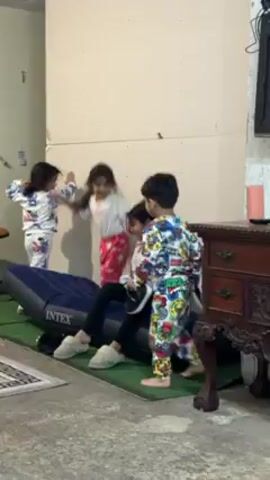 Four young children are indoors, likely in a playroom or basement. A girl in white and blue pajamas jumps repeatedly on a deflated blue Intex air mattress resting on a green carpet. Another girl in a white shirt and red pants sits on the mattress. A third girl, dressed in black pants and a white shirt, sits with her legs stretched out, looking towards the other children. A young boy in colorful pajamas sits next to her, looking at the camera. A small dark wooden table with ornate carvings sits to the right of the group. The walls are cream-colored and appear to be unfinished. A TV is mounted on the wall in the background. A rug sits on the bare concrete floor in the foreground.