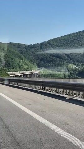 A view from a vehicle moving along a highway. In the foreground is the asphalt road with a white line marker. A metal guardrail runs along the side of the road. In the background, a damaged bridge is visible, with sections appearing to be collapsed. The bridge is situated in a valley with lush green, tree-covered mountains rising behind it. The sky is clear and blue with a few wisps of white clouds. There are no people or animals visible in the frame.