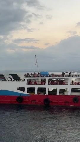 The video shows a ferryboat traveling on the water. The ferryboat is red and white with blue accents. There are people on the upper deck of the ferryboat. The sky is cloudy and the water appears choppy. The ferryboat is moving from left to right across the screen.