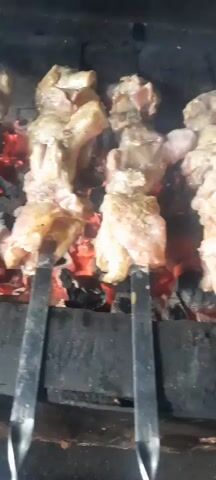 Four metal skewers, each holding several pieces of raw meat, are positioned over a bed of glowing red embers in a charcoal grill. The meat appears to be chicken, with some pieces still attached to bones. Smoke rises from the grill. The scene is dimly lit, with the primary light source coming from the glowing coals.