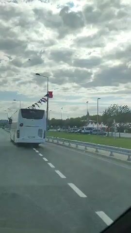 A white bus is driving on a highway. The bus is in the right lane. There are other vehicles on the highway. There are trees and buildings in the background. The sky is cloudy. There are flags strung across a pole on the side of the highway.