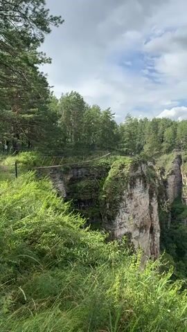 The video pans from left to right, revealing a steep cliff face covered in greenery, with a path winding along the top. The path is bordered by a simple wooden fence. Beyond the cliff, a wide, calm river flows through a lush green forest that stretches to the horizon. The sky is cloudy, with patches of blue showing through. In the distance, on the river, there are two small boats, one yellow and one red.