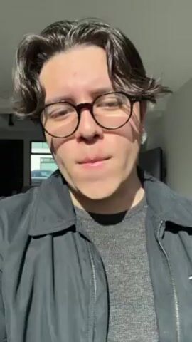 A young man with round glasses is seen from the chest up. He is wearing a gray sweater and a black zip-up jacket with a Tommy Hilfiger logo. He looks directly at the camera, touches his hair, and then looks away. The scene is brightly lit, likely from natural light.