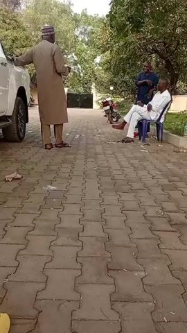 The video shows three men outside on a brick-paved driveway. One man wearing a brown tunic, matching pants, and a brimless cap stands with his hand on the side of a white pickup truck. The second man, wearing a dark blue shirt and black pants, stands near a man sitting on a light blue plastic chair. The man sitting on the chair is wearing white clothing. The scene appears to be a driveway in front of a building obscured by trees. A motorcycle is parked in the background. The men are in discussion and appear to be at ease.