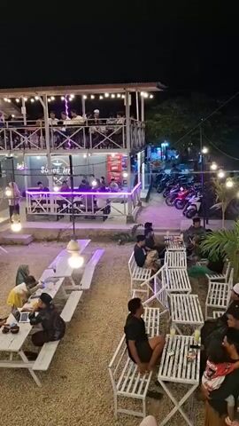 The video shows an outdoor seating area of a restaurant or cafe at night. Several people are seated at picnic tables and benches. Some tables have white tops and white benches. There are also several small bamboo huts with thatched roofs, each containing tables and people. In the background, there is a two-story building with lights and people on both levels, appearing to be part of the establishment. String lights are hung throughout the outdoor area and around the two-story building. There are also motorcycles parked in the background. The ground is covered in gravel. The overall ambiance is casual and social.