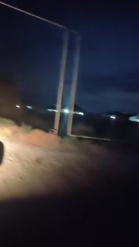 A first-person perspective video shows a dark, nighttime scene. The view is from inside a vehicle, looking out at a dirt road on the left side. The road is illuminated by headlights. In the distance, there are several buildings with lights on. The sky is dark blue with some clouds. There are two vertical poles in the foreground, obscuring part of the view. The overall scene appears to be rural.