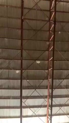 The camera is pointed up at the roof of a large, indoor structure. The roof is made of corrugated metal and is supported by metal beams and crossbeams. Light is coming through the roof, and there are a few light fixtures visible. The camera pans down to show a white wall and a person sitting in the corner. The person is wearing dark clothing and has their legs crossed. There is some background noise.