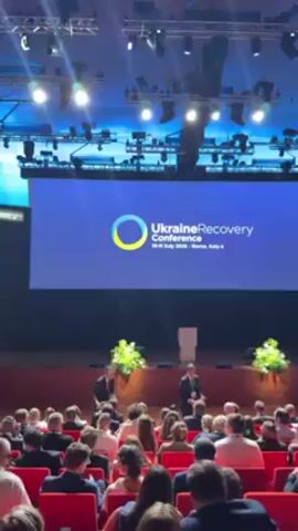 The video captures a view from the audience during a Ukraine Recovery Conference. On stage, two men stand at a podium in front of a large screen displaying the conference logo. The screen shows the "Ukraine Recovery Conference" title and the dates "10-11 July 2023 - Rome, Italy." The conference is taking place in a large auditorium, filled with seated attendees facing the stage. The auditorium features red seats, a wooden stage, and various stage lights overhead. The attendees are diverse and dressed in business attire.