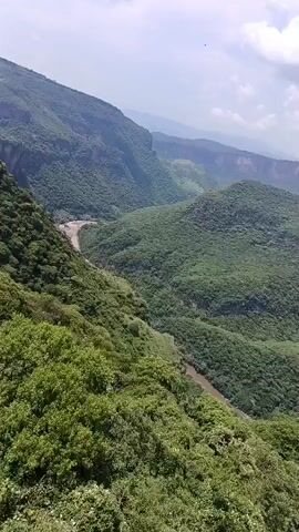The video displays a panoramic view of a lush green mountainous landscape. The camera pans across a deep canyon with a river flowing through its bottom. The sides of the canyon are heavily forested with dense vegetation. In the distance, a hazy skyline suggests the presence of a city or town. The sky is partly cloudy with a bright, daytime sun. The overall impression is one of natural beauty and vastness.