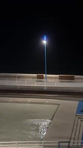 This is a vertical video taken at night of an outdoor area that appears to be a public space, possibly near a waterfront or pool. The lighting is dim, with several tall, blue-tipped lampposts illuminating the scene. In the foreground, there is a pool of water with visible ripples, suggesting it is either a swimming pool or a water feature. Adjacent to the pool is a blue-tiled area with some scattered colorful play equipment. Further back, a paved walkway is lined with benches. The background is dark, with the ocean or a large body of water visible behind the walkway. The video pans slightly to the right, revealing more of the walkway and another lamppost.