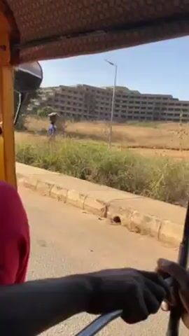The video shows a person driving a yellow tuk-tuk on a paved road in what appears to be a rural or semi-urban area. A woman carrying something on her head walks along a dirt path parallel to the road. In the background, there is a large, unfinished or dilapidated building that appears to be multi-story. The vegetation around the building is dry and brownish. The sky is clear and blue. A sign for a resort and accommodation is briefly visible.