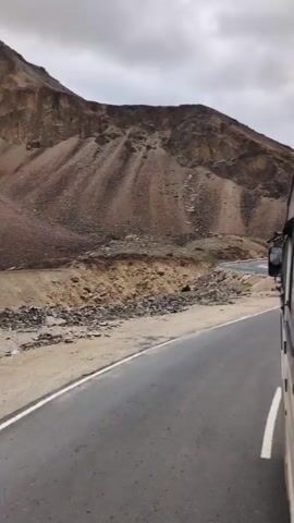 The video captures a view from a moving bus on a winding mountain road. The road is paved and has white lines marking the lanes. The landscape is rugged and mountainous with dry, rocky slopes. There is some sparse vegetation on the sides of the road. The sky is overcast with clouds. The bus is seen from the side, with its windows visible. The bus is navigating a curve in the road, with a metal guardrail along the outer edge. The scene appears to be in a remote, high-altitude region.