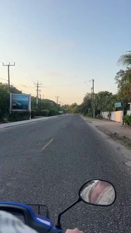 The video shows a first-person perspective of someone riding a blue scooter down a paved road. The road is mostly straight and has a single yellow line marking the center. On either side of the road, there are trees and vegetation. Power lines run along the side of the road supported by utility poles. A billboard is visible on the left side of the road. The sky is light blue with some hints of orange at the horizon, suggesting it is either early morning or late afternoon. The scooter's side mirror shows the reflection of the rider's clothing, which appears to be a light-colored, textured shirt. The rider is wearing a blue helmet, partly visible at the top of the frame.