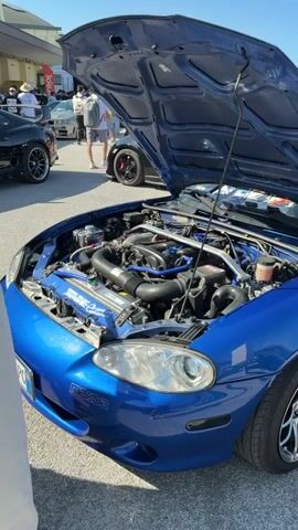 The video shows a blue Mazda Miata with its hood open, revealing the engine. The car is parked outdoors, likely at a car show or exhibition, given the presence of other cars in the background and people milling around. The scene appears to be well-lit, suggesting it is daytime with clear weather, possibly a sunny afternoon.

The engine bay is customized with blue accents on hoses and a strut brace, indicating performance modifications. There is a prominent "Mazda" logo on the front of the car. Visible text on the engine cover includes "MADE IN JAPAN" and what appears to be a Japanese flag graphic, suggesting parts sourced from Japan. Another sticker on the radiator says "MONDO racing."

In the background, several other vehicles are visible. To the left, a black sports car with a spoiler is parked, and further back, a light-colored car, possibly a Nissan Skyline, is also present. Another sports car with red brake calipers is parked behind the Miata. People are seen walking around the vehicles, observing them. The overall atmosphere seems to be one of appreciation for automobiles, typical of a car enthusiast event. The location context provided is Cesena, Italy.
