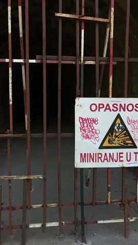 This media file shows the entrance to a dark tunnel, possibly a bunker or part of a military installation. The entrance is blocked by a rusty metal gate with vertical bars, some of which have red and white painted stripes. To the right of the gate, a white rectangular sign is visible. The sign has a yellow triangular warning symbol with a black silhouette of a person and a bomb, indicating danger. Below the symbol, in large red letters, is the word "OPASAN", which translates to "DANGER" in Serbian. Further down on the sign, in smaller red letters, is the word "MINIRANJE", meaning "MINING" or "MINEFIELD". Red graffiti is spray-painted over the warning symbol and the word "OPASAN".

The tunnel itself recedes into darkness, with a concrete or asphalt pathway visible on the floor. The walls of the tunnel appear to be constructed from stone. Two small, faint lights can be seen in the distance within the tunnel. The overall impression is one of a forbidden and potentially hazardous location. The presence of the warning sign and the locked gate suggests that entry is prohibited due to safety concerns, likely related to landmines. The scene evokes a sense of mystery and caution. The time of day and weather are not discernible from the image.