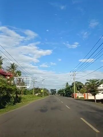 The video shows a POV driving on a paved road. The sky is bright blue with scattered white clouds. On either side of the road are telephone poles with wires running overhead, houses, trees, and green foliage. There are several signs visible along the road. One sign on the left is a large billboard. A yellow diamond-shaped sign with a black arrow pointing left is visible on the left side of the road. The road is marked with white lines. The time stamp indicates the video is from 00:00 to 00:04.