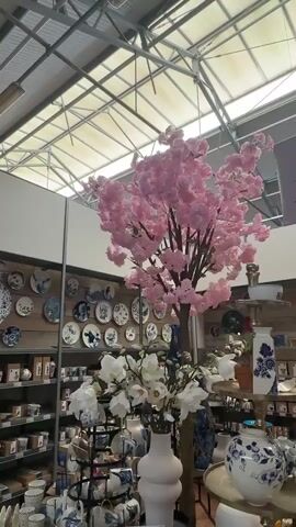 The video pans across a retail store, likely a home goods or garden center, with a focus on decorative items. The foreground features a large artificial pink cherry blossom tree, prominently placed. To the left and right of the tree are displays of ceramics, including plates with blue and white designs reminiscent of Delftware, and various vases and mugs. White artificial flowers in a pink vase are also visible in the lower left. The background shows shelving units stocked with more merchandise. The store has a high ceiling with exposed industrial-style lighting and a translucent roof structure, allowing natural light to filter in. The overall impression is that of a well-lit retail environment showcasing decorative home accents and possibly garden-related items.