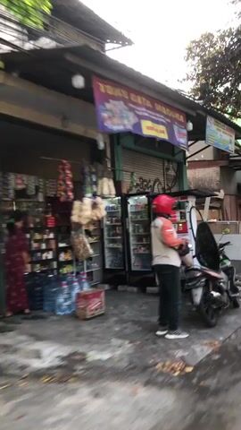 The media file shows a street scene in Candi, East Java. The primary subject is a person wearing a red helmet, an orange and white long-sleeved shirt, dark pants, and sneakers. They are standing next to a motorcycle, with a bag slung over their shoulder. The person appears to be looking towards a storefront.

The scene is set in front of a small shop or convenience store, identified by the banner overhead that reads "TOKO GITA MADURA". The store has shelves stocked with various goods, including what appear to be snacks or packaged food items. A refrigerator unit is visible, filled with products. Several bottles of water and a boxed item are placed on the ground near the entrance.

The motorcycle is a prominent object in the foreground, parked close to the person and the storefront. It's a red and black scooter-style motorcycle.

The lighting suggests it is daytime, possibly late morning or afternoon, with no strong indications of specific weather conditions. The environment appears to be urban or semi-urban, with a paved ground surface and a shop building.

The overall mood is neutral, depicting a common everyday scene of someone at a local store. There is no strong indication of any particular emotion.

Visible text on the banner includes "TOKO GITA MADURA", and below that, some Indonesian text which seems to indicate opening hours or services, with "BUKA 24 JAM" clearly legible, meaning "Open 24 Hours". There is also text on some of the products inside the store and on the box on the ground.